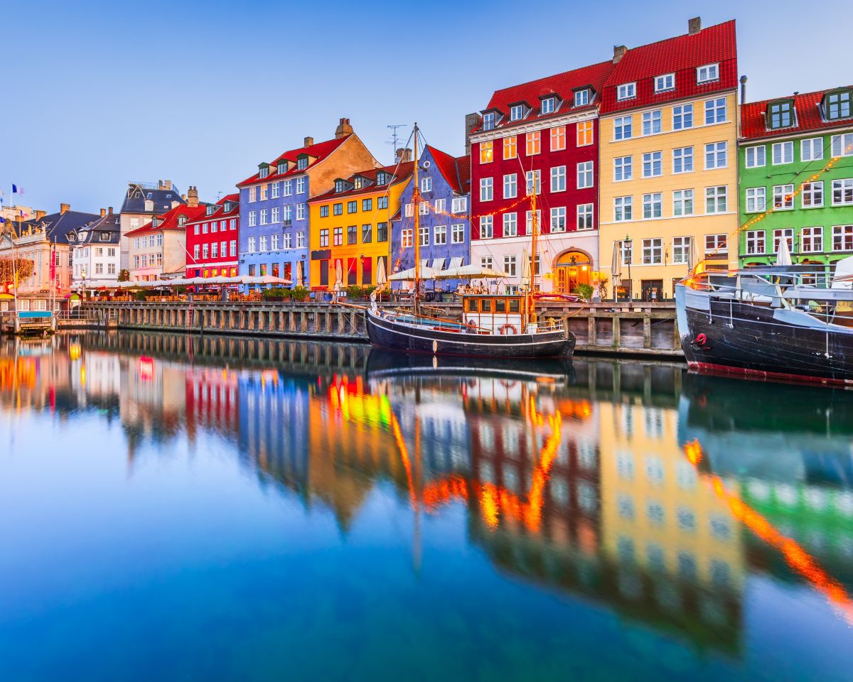 Colorful historic buildings lining Nyhavn harbor in Copenhagen, Denmark, with boats reflected in calm water at dusk.